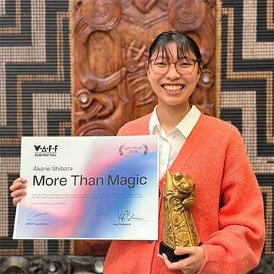 Award winner Akane Shibata smiling and holding a certificate reading "More Than Magic" and a golden trophy statue, standing in front of a carved wooden panel backdrop at the VAFF (presumably a film festival).