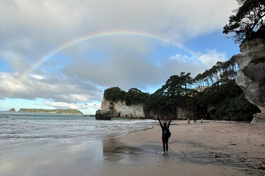 Sydney with raised arms on the beach on a cloudy day, a rainbow is shining through the clouds