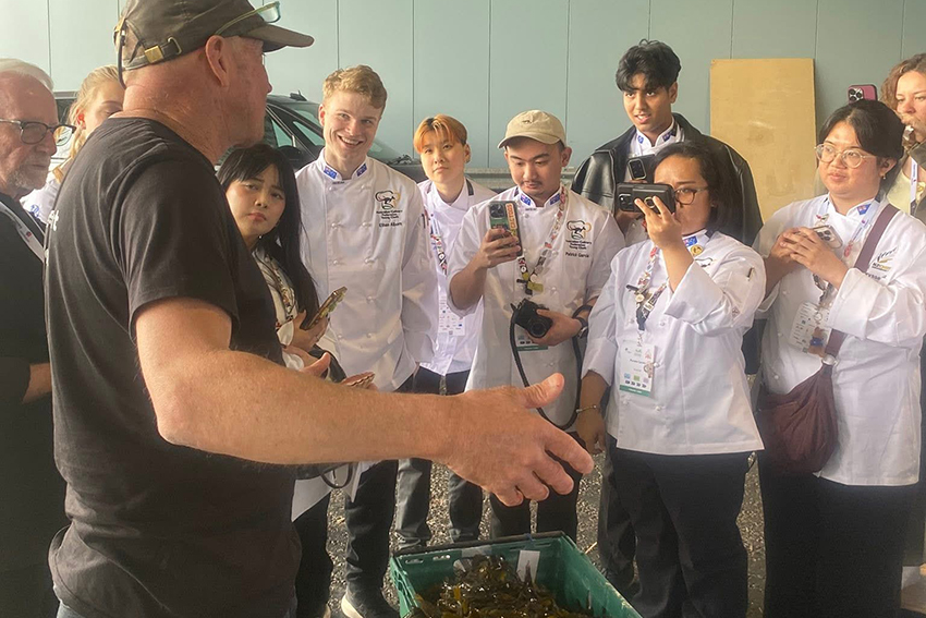A man in a black T-shirt presenting a pastic crate of scallops to a group of student's in whites, some of whom are recording the conversation on their phones.