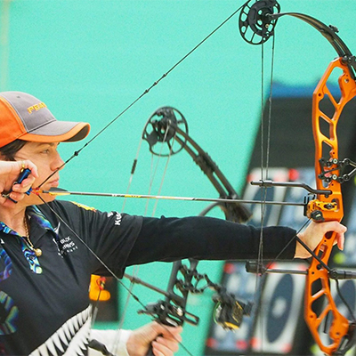 Compound archer in a Black Arrows New Zealand shirt draws an orange bow at an indoor range, with targets behind.
