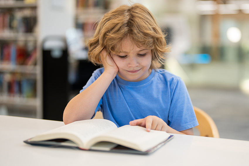 A child of perhaps 7 sitting in a library reading a book at a desk with a smile on his face.