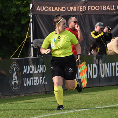 Assistant referee in a fluoro yellow New Zealand Football kit runs the touchline holding a flag at an Auckland United match.