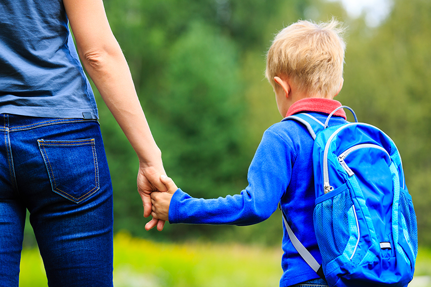 Generic photo of a child with a schoolbag holding his mother's hand, shot from behind with faces not visible.
