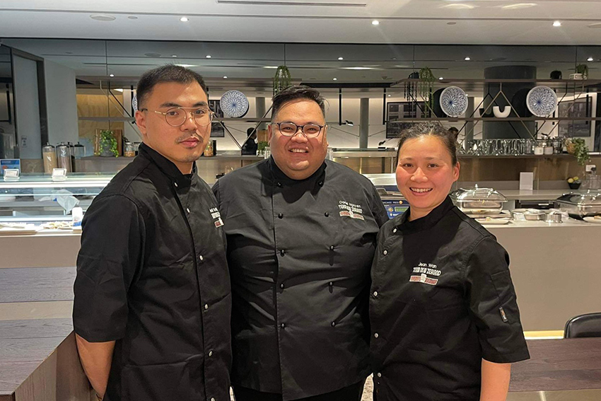 Three members of the team standing in chef's clothes in front of a buffet.