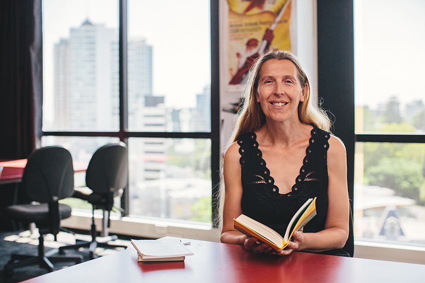 Siobhan holding a book in a classroom, smiling at the camera, with a view over Auckland through windows behind her.