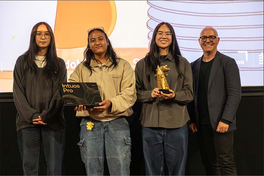 Four people standing together on a stage at an awards ceremony. The second person from the left holds a Wacom Intuos Pro box, and the third holds a golden trophy statue. A presentation screen is visible in the background.