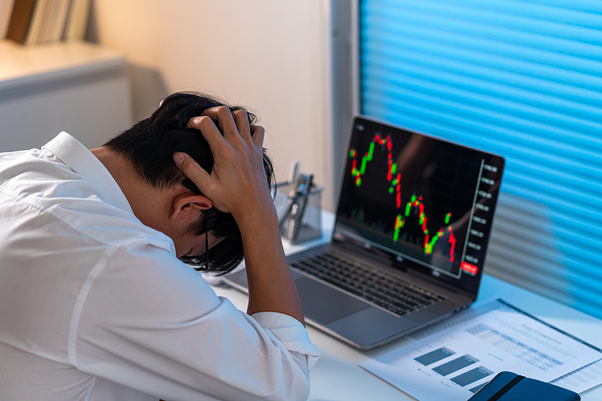investor regret A man in a shirt with his head in his hands, sitting in front of a laptop showing declining stock prices..