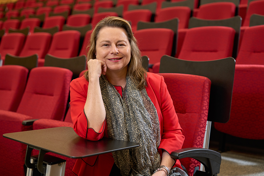 Brigitte sitting in a lecture theatre, surrounded by empty red seats. she has her elbow leaning on the seat desk and smiling at the camera.