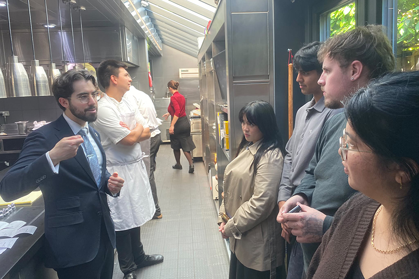 In a professional kitchen, a man in a suit is instructing the four students, who are listening intently.