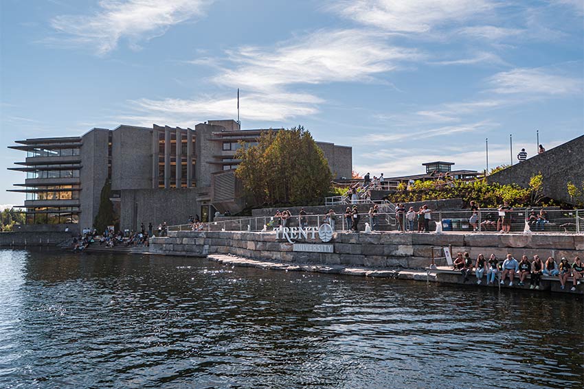 Trent University's waterfront building seen from across the river, with students gathered along the stone terrace on a sunny day.