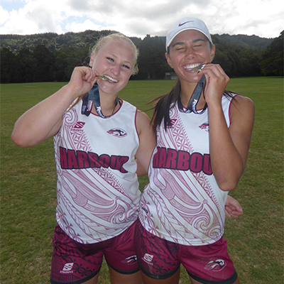 Two touch players in Harbour singlets smile as they bite their medals on a grass field.