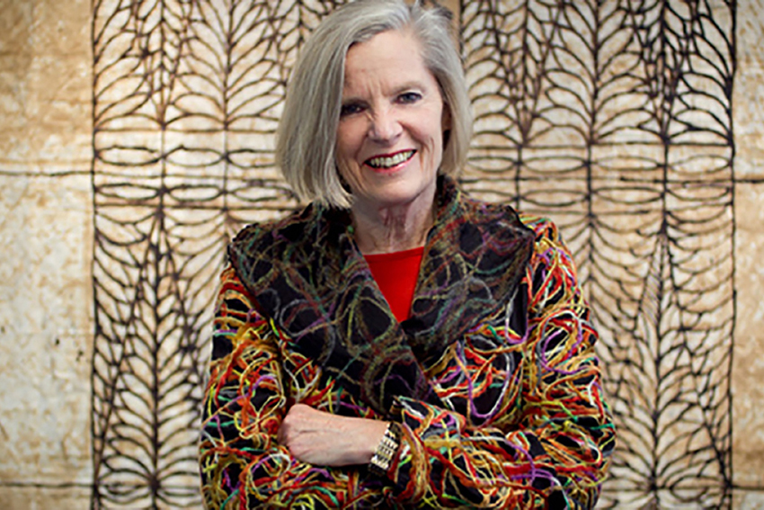 Dame Judy McGregor standing arms crossed and smiling in front of a Pacific tapestry