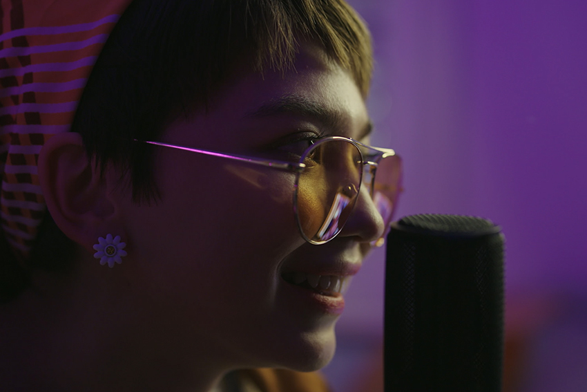 Teenage woman with glasses speaking into a microphone from a side angle.