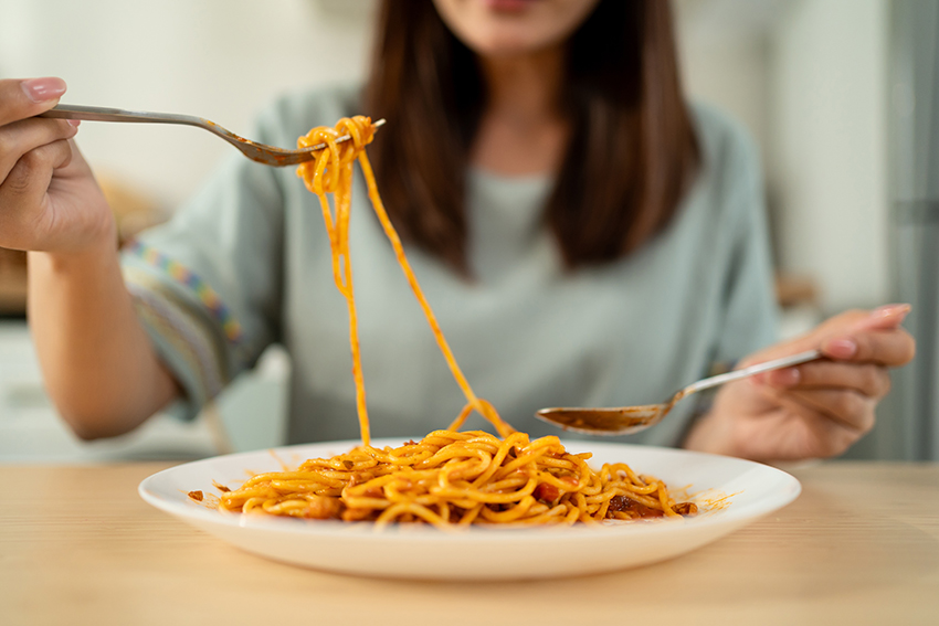 A woman eating pasta with a fork and spoon. Depth of focus is on the pasta with the woman slightly blured behind.