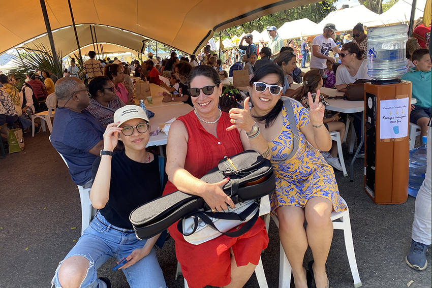 Dianne, Tracy and Dolby smiling at the camera amongst festival goers people sitting at tables under large shade sails.