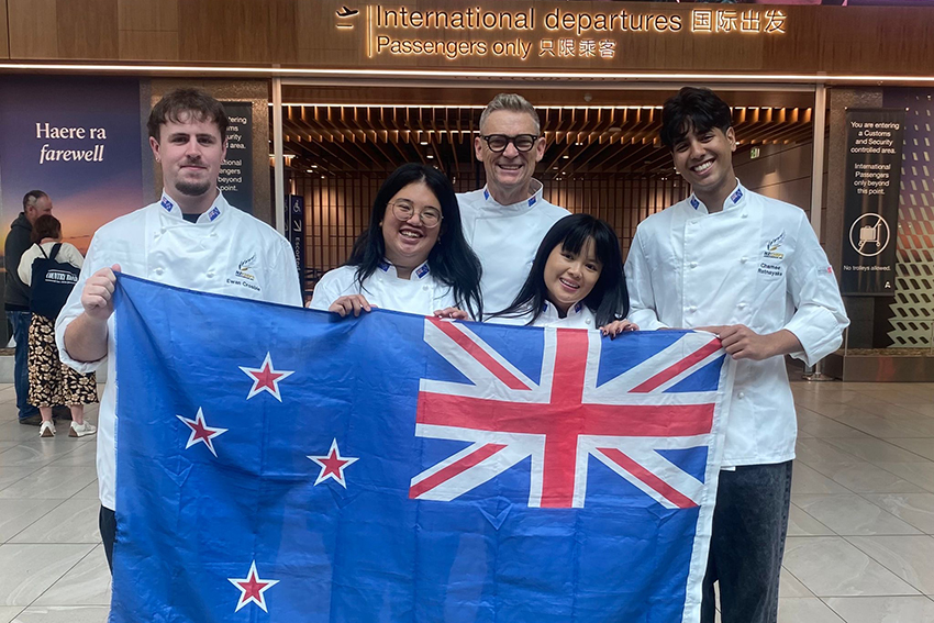 The students and Geoff holding a NZ flag and standing in front of an airport departures sign.