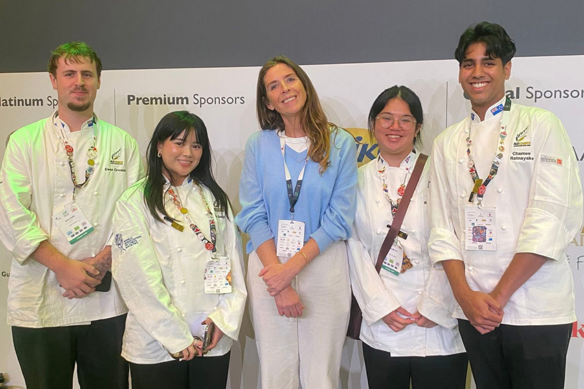 A woman in a bluejumper and four students in chef whites, alined up and ll smiling.