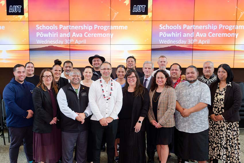 A group of people involved with the AUT and Schools partnership is standing in front of a lit-up screen