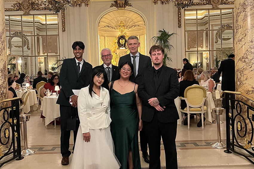 The students standing with Geoff and Tony, dressed up and in a room with gold trim and fancy tables and diners.