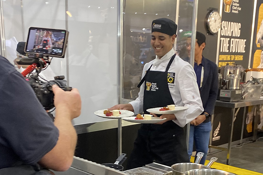 A young man in chef whites and competition apron holds three plates of dessert. He is smiling and being filmed.