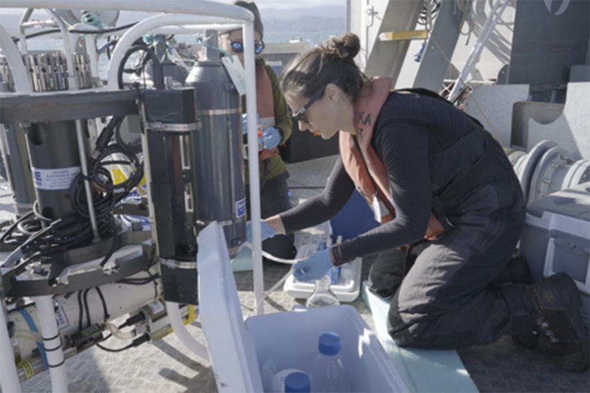 Marta crouched down wearing a life jacket, sunglasses and gloves n the deck of a boat that is crowded with equipment that she is using.