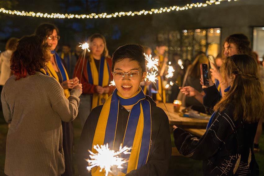 A student wearing a blue and yellow scarf holds sparklers at an evening outdoor gathering, surrounded by others also holding sparklers under string lights.