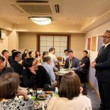 Person in a suit speaking to a group seated around dining tables in a restaurant room.