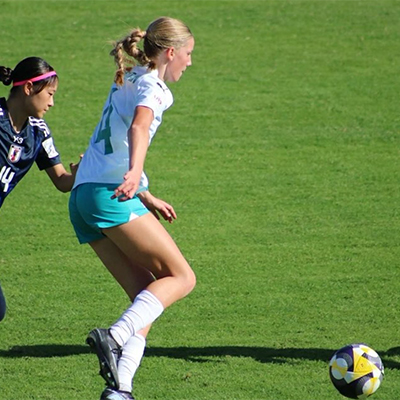 Football player in a white and teal kit dribbles the ball while closely marked by a Japan player in navy, number 14.
