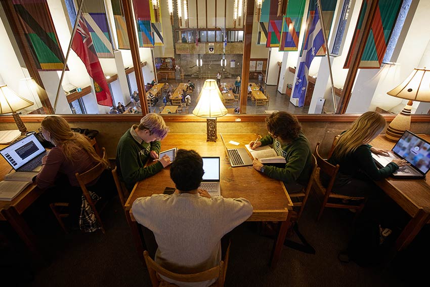 An overhead view of students studying at wooden library tables beneath tall windows, with colourful banners hanging on the walls.