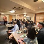 Person speaking at the front of a restaurant dining room while attendees seated at tables listen.