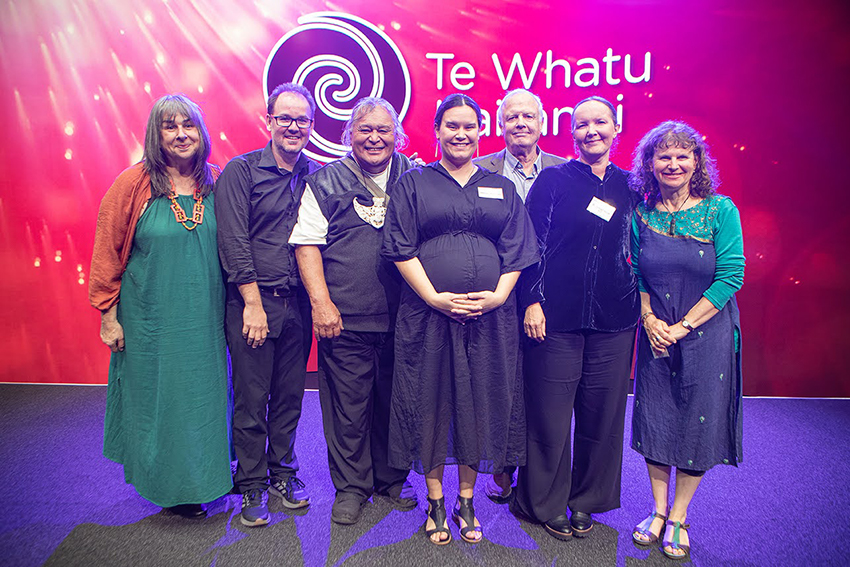 Seven people dressed up and standing smiling with Te Whatu Kairangai branding behind them.