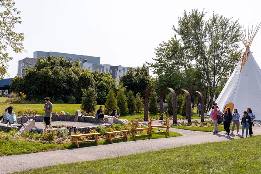 Students walking through a landscaped garden area with a teepee, metal arch sculptures, and wooden benches, with a university building in the background.