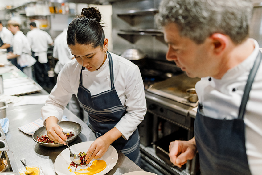 Alex in chef whites and an apron places ingredients on a white plate in a busy kitchen, with Gavin watching.