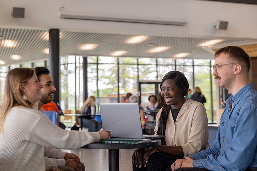 Four university students chatting and smiling around a laptop at a table in a bright, glass-walled common area.