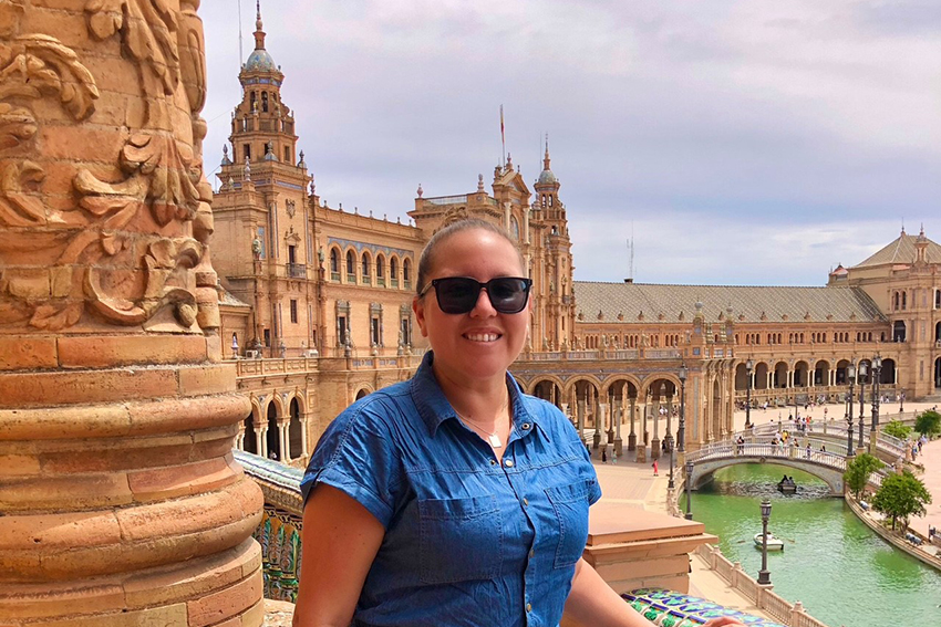 A person with sunglasses on standing in front of a magnificent old orange building with a canal in front.